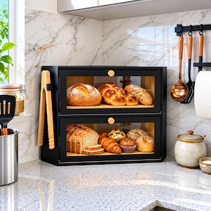 May include: A two-tier black bread box with clear glass doors and light wood accents. The top shelf holds a round loaf, a braided loaf, and a baguette. The bottom shelf contains sliced bread, a croissant, muffins, and a bagel. Wooden tongs hang on the side.