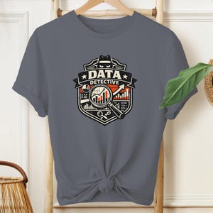 May include: A charcoal gray t-shirt featuring a graphic emblem with the text DATA DETECTIVE. The design includes a magnifying glass over a bar chart, a pie chart, and a silhouette of a person wearing a fedora hat, all rendered in black, white, and orange.