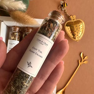 May include: A clear glass test tube filled with loose leaf tea, featuring a white label with the text Calm & Chaos, Erst Tee, Dann Drama. Beside it lies a gold heart-shaped tea infuser with a bee charm and a gold spoon with a leaf handle.