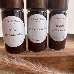 May include: Three clear glass jars filled with dark brown ground coffee, each topped with a dark brown wood-grain lid. Each jar features a white oval label with the text Calm & Chaos Studio and unique phrases: KEIN SMALLTALK, ERST KAFFEE, and DANN VIELLEICHT.