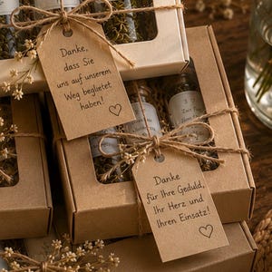 May include: A collection of rectangular cardboard gift boxes with clear windows, each containing glass vials filled with dried herbs. The boxes are tied with twine and decorated with small dried flowers and kraft paper tags featuring German text.