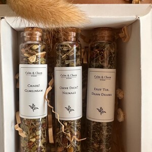 May include: Three clear glass test tube bottles with cork stoppers filled with loose leaf tea blends. Each bottle features a white label with the text Calm & Chaos, followed by the phrases Chaos? Gemeinsam, Ohne Dich? Niemals, or Erst Tee. Dann Drama.