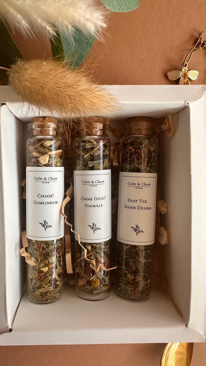 May include: Three clear glass test tube bottles with cork stoppers filled with loose leaf tea blends. Each bottle features a white label with the text Calm & Chaos, followed by the phrases Chaos? Gemeinsam, Ohne Dich? Niemals, or Erst Tee. Dann Drama.