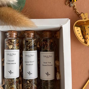 May include: Three glass test tubes with cork stoppers filled with loose leaf tea, each featuring a white label with the text Calm & Chaos, followed by the phrases Chaos? Gemeinsam, Ohne Dich? Niemals, and Erst Tee, Dann Drama, inside a white box.