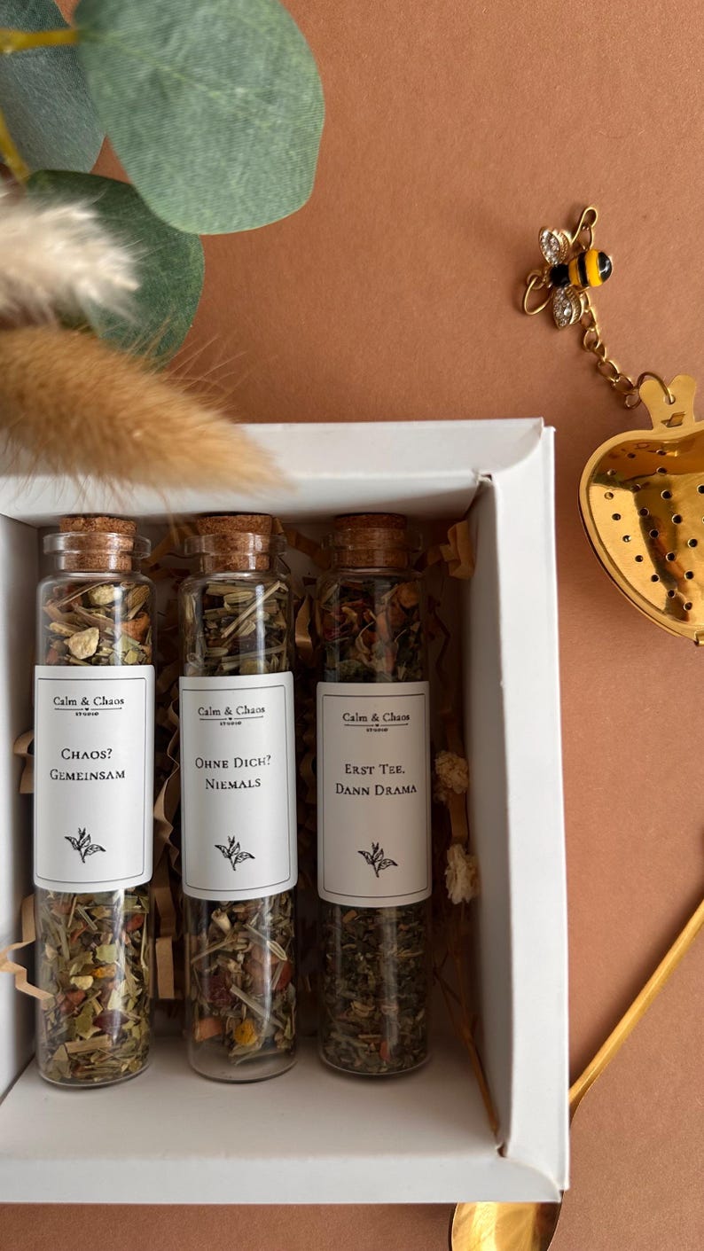 May include: Three glass test tubes with cork stoppers filled with loose leaf tea, each featuring a white label with the text Calm & Chaos, followed by the phrases Chaos? Gemeinsam, Ohne Dich? Niemals, and Erst Tee, Dann Drama, inside a white box.