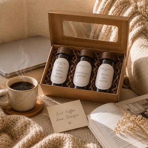 Pode incluir: Uma caixa de presente de papelão com uma janela transparente contendo três potes de vidro com tampas de madeira escura e etiquetas brancas com os dizeres Erst Kaffee, dann Therapie, Emotional geröstet e Stress neu starten, ao lado de uma caneca.