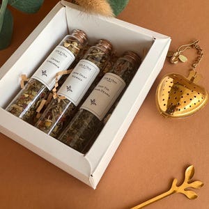 May include: A white gift box containing three glass test tubes filled with loose leaf tea, each with a cork stopper and a label reading "Calm & Chaos". A gold-colored heart-shaped tea infuser and a gold-colored spoon with a leaf-shaped handle.