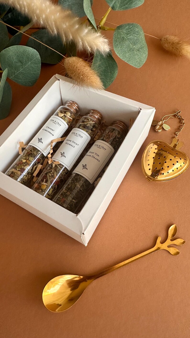 May include: A white gift box containing three glass test tubes filled with loose leaf tea, each with a cork stopper and a label reading "Calm & Chaos". A gold-colored heart-shaped tea infuser and a gold-colored spoon with a leaf-shaped handle.