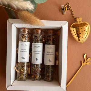 May include: A white gift box containing three glass test tubes filled with loose leaf tea, each with a cork stopper and a white label reading Calm & Chaos, followed by the phrases Chaos? Gemeinsam, Ohne Dich? Niemals, and Erst Tee Dann Drama.