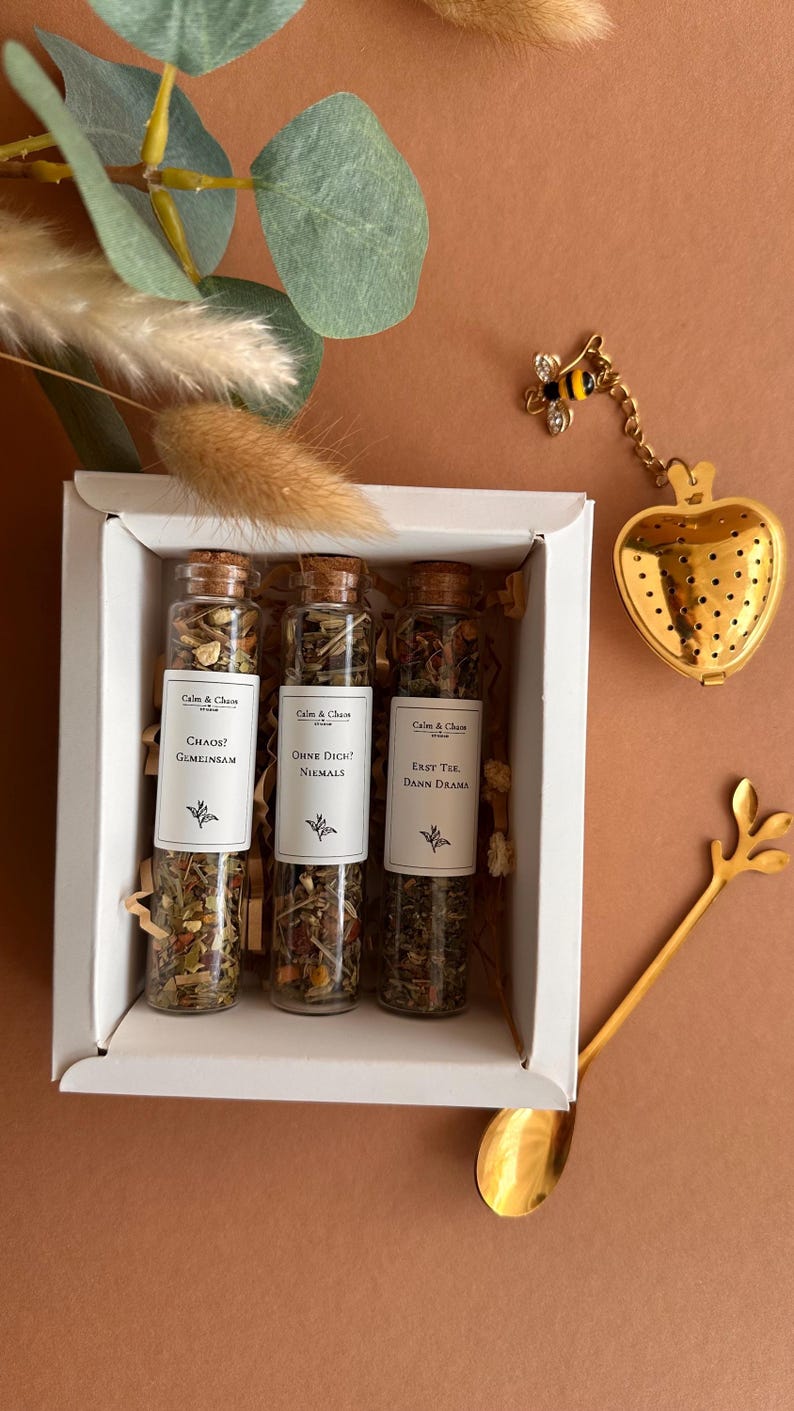 May include: A white gift box containing three glass test tubes filled with loose leaf tea, each with a cork stopper and a white label reading Calm & Chaos, followed by the phrases Chaos? Gemeinsam, Ohne Dich? Niemals, and Erst Tee Dann Drama.