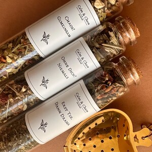 May include: Three glass test tubes filled with loose leaf tea, each sealed with a cork stopper and a white label featuring the text Calm & Chaos, plus German phrases. A gold-colored heart-shaped tea infuser with a bee charm sits nearby.