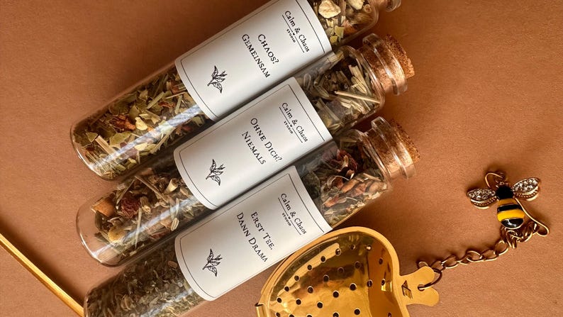 May include: Three glass test tubes filled with loose leaf tea, each sealed with a cork stopper and a white label featuring the text Calm & Chaos, plus German phrases. A gold-colored heart-shaped tea infuser with a bee charm sits nearby.