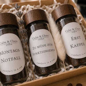 Könnte beinhalten: Drei Glasbehälter mit dunklen Holzdeckeln gefüllt mit Kaffeepulver, eingebettet in eine Pappschachtel mit Papierfüllmaterial. Jedes Glas hat ein weißes ovales Etikett mit dem Text Calm & Chaos Studio, comfort in a cup und Sprüchen.