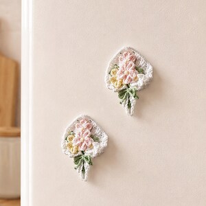 May include: Two handmade crochet flower magnets featuring small pink and yellow blossoms with green leaves, arranged in a white crocheted bouquet shape. Each piece has a textured, intricate yarn construction and is designed to adhere to surfaces.