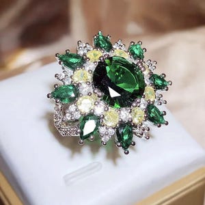 Puede incluir: Un anillo de tono plateado con una gran piedra central redonda de color verde oscuro, rodeada por un halo de piedras verdes más pequeñas en forma de pera, piedras amarillas redondas y diminutas piedras blancas transparentes en un diseño floral.