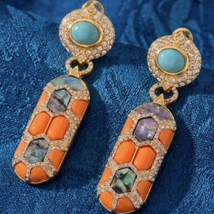 Könnte beinhalten: Ein Paar goldfarbene Ohrhänger mit einem runden Oberteil, das einen zentralen türkisfarbenen Cabochon und klare Kristalle aufweist. Der längliche untere Teil zeigt ein geometrisches Muster aus orangefarbenen Steinen und Perlmutt-Einlagen.