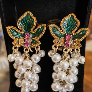 Può includere: Un paio di orecchini pendenti con un design a foglia smaltata verde, accentuato da piccoli strass color ambra e due strass rosa al centro. Sotto la foglia, grappoli di perle finte bianche pendono da maglie in metallo color oro.