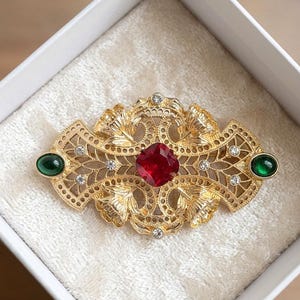 Può includere: Una spilla in metallo color oro con un intricato design in filigrana. Il pezzo è centrato da una pietra preziosa rossa a taglio quadrato, affiancata da due cabochon verdi ovali alle estremità e accentuata da piccoli strass trasparenti.