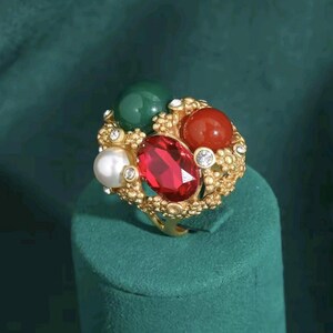 May include: A gold-toned ring featuring a textured, organic-style band adorned with a central oval-cut red gemstone, a round green bead, a round red bead, a white pearl, and several small clear rhinestones embedded throughout the metal setting.