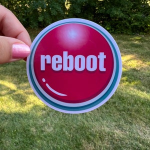 May include: A round sticker with the word "reboot" in white, set against a red background. The design includes a white and teal border, with a bubble-like accent. The sticker is held against a backdrop of green grass.
