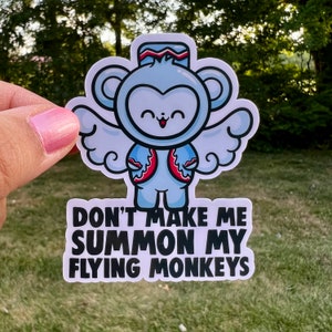 May include: A white sticker with a cartoon monkey wearing a red and blue hat and wings. The monkey has a happy expression and is surrounded by a cloud-like design. The text "Don't make me summon my flying monkeys" is written in black below the monkey.