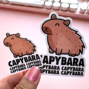 Pode incluir: Dois adesivos de capivara marrons com contornos pretos. O adesivo maior tem a palavra "CAPYBARA" repetida quatro vezes em texto preto. O adesivo menor tem a palavra "CAPYBARA" repetida quatro vezes em texto preto.