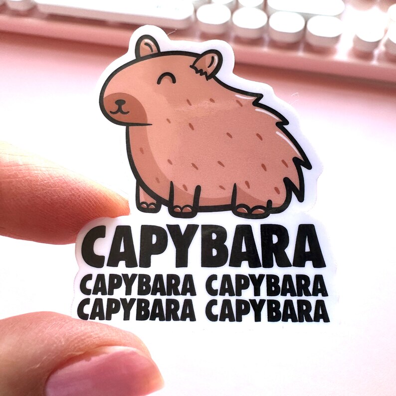 Pode incluir: Um adesivo branco com uma ilustra&ccedil;&atilde;o de capivara marrom de desenho animado. O adesivo tem a palavra "CAPYBARA" escrita em texto preto quatro vezes.