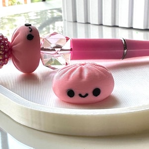May include: A pink pen featuring a decorative topper shaped like a round, pleated bun with a smiling face, accompanied by a separate, matching pink bun-shaped object with a black-painted smiling face and two black circular eyes.
