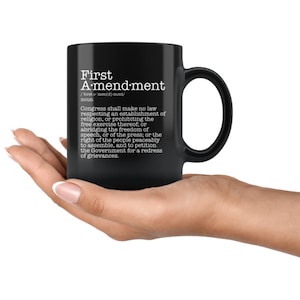 May include: Black coffee mug with white text that reads "First Amendment" and the definition of the First Amendment to the United States Constitution.