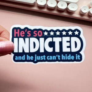 May include: A white sticker with blue and red text that reads "He's so INDICTED and he just can't hide it". The sticker has five stars above the word "INDICTED".