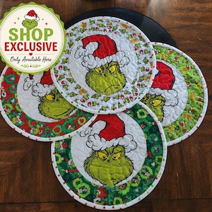 May include: Three round quilted fabric mats featuring a green character wearing a red and white Santa hat. The mats have a central image of the character surrounded by a border of green wreaths or a pattern of small green characters on a white background.