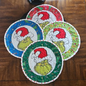 May include: Four round quilted placemats featuring a green Grinch character wearing a red Santa hat. The placemats have a white background with a green, red, and blue patterned border.