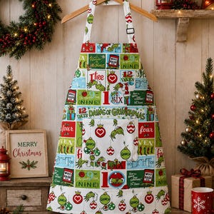 May include: A white apron featuring a colorful grid pattern with illustrations of the Grinch, Christmas ornaments, and text reading 12 Days of Grinchmas. The design includes red, green, and blue accents, a central pocket, and an adjustable neck strap.