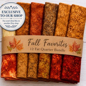 May include: A bundle of 12 folded fabric fat quarters in various shades of orange, red, brown, and gold with assorted patterns. A brown paper band wraps around the center with the text Fall Favorites 12 Fat Quarter Bundle and maple leaf graphics.