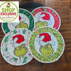 May include: Four circular quilted fabric mats featuring a central illustration of a green character wearing a red Santa hat. Each mat has a distinct patterned border in red, green, or white, decorated with holiday-themed motifs and festive imagery.