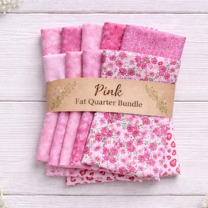 May include: A bundle of pink fat quarters, featuring various shades and patterns, including floral and heart prints. The bundle is wrapped with a brown paper band that reads "Pink Fat Quarter Bundle". The fabrics are arranged on a white wooden surface.