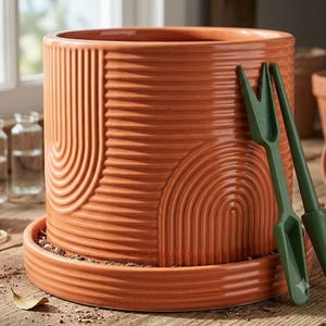 May include: A cylindrical terracotta-colored ceramic planter with a matching saucer, featuring a textured surface with embossed vertical and arched line patterns. Two dark green plastic gardening tools are leaning against the side.