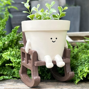 May include: A cream-colored ceramic planter shaped like a pot with a simple black smiley face, featuring small arms and legs, seated in a brown wooden-style rocking chair, containing a small green plant with rounded leaves.