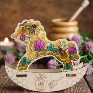 May include: A wooden rocking horse ornament featuring a cross-stitch pattern of yellow honeycomb, bees, pink clover flowers, and green leaves. The base is engraved with a honey jar labeled Meg and a honey dipper, all within a honeycomb pattern.