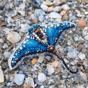 May include: A stingray-shaped brooch featuring a gradient of blue and dark blue thread embroidery. The piece is embellished with white pearls, metallic gold beads, and larger bronze-colored faceted beads along the body and tail.