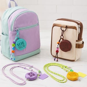 May include: A lavender and light blue backpack and a beige and brown rectangular bag, each featuring a circular coin pouch with the word science embossed on it and a colorful beaded charm. Two additional circular coin pouches with lanyards.