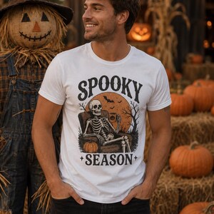 May include: A white short-sleeved t-shirt featuring a graphic print of a skeleton sitting in an armchair, holding a mug, with a large orange moon, bats, and trees in the background. The text SPOOKY SEASON is printed in black above and below.