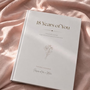 May include: A white hardcover journal with gold text that reads 18 Years of You, A keepsake journal of growth, memories, and moments, Your story, your journey, Preserved for a lifetime, featuring a delicate line drawing of a flower in the center.