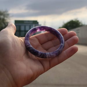May include: A smooth, circular bangle bracelet crafted from polished amethyst stone. The bracelet features deep purple hues mixed with translucent white bands and natural mineral inclusions throughout the solid, rounded stone structure.