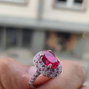 Puede incluir: Un anillo de tono plateado con una gran piedra central de color rojo vibrante y corte ovalado. El engaste está rodeado por un halo de piedras más pequeñas de corte redondo en rojo y blanco transparente, con piedras adicionales en el aro.