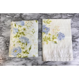 May include: A folded white pillowcase and a flat white pillowcase, both featuring a floral pattern of light blue roses and green stems. The flat pillowcase includes a decorative white eyelet lace border along the bottom edge.