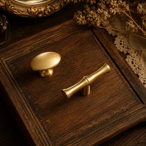 May include: Two brass cabinet knobs with a brushed gold finish. One knob is oval-shaped with a smooth, rounded top, while the other is a T-bar style handle with a central cylindrical post and flared ends, both designed for furniture hardware.