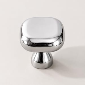 May include: A polished chrome cabinet knob featuring a square head with rounded corners and a smooth, reflective surface. The base is circular and tapers slightly toward the center, providing a sleek, metallic finish for furniture hardware.