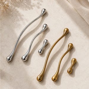 May include: A collection of six metal cabinet pulls in varying lengths and finishes. Three pulls feature a polished silver finish, while three feature a brushed gold finish. Each pull has a slender, curved, organic shape with rounded ends.