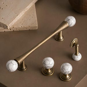 May include: A set of cabinet hardware including one long bar handle and three round knobs. Each piece features a brushed antique brass finish and white spherical accents with a grey marble-like veined pattern throughout the stone surface.